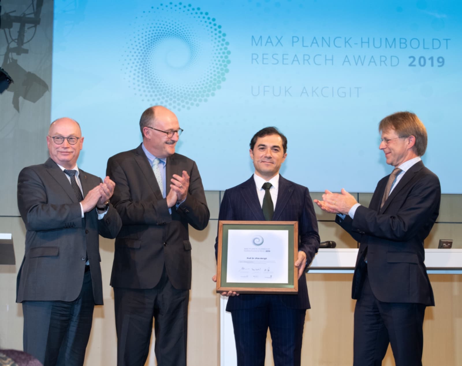 Ufuk Akcigit on stage at the Berlin Science Week award ceremony, holding a framed Max Planck–Humboldt Research Award certificate, alongside three representatives of the Max Planck Society and the Humboldt Foundation, November 2019.